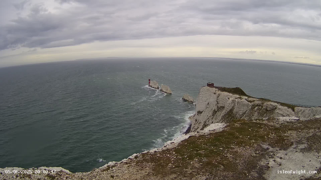 A view from a webcam shows rugged white chalk cliffs extending into the sea under a cloudy sky. A small lighthouse is situated on the rocks in the ocean, with a red and white striped pattern. A building, likely a shelter or observation point, is located near the edge of the cliffs on the right side. The water below is choppy, and the overall scene has a moody, overcast atmosphere. The timestamp at the bottom left indicates the image was captured on May 6, 2025, at 8:00:42 PM. The website "isleofwight.com" is visible in the bottom right corner.