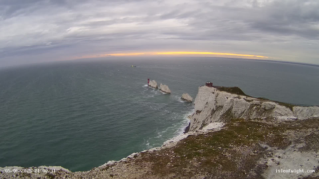 A view from a webcam showing white cliffs along a rugged coastline with a small building or lighthouse at the edge. The ocean extends into the distance under a cloudy sky with a hint of sunset light on the horizon. A boat is visible near the cliffs, and the overall scene is peaceful and natural. The timestamp indicates the date and time as May 6, 2023, at 21:00:31.