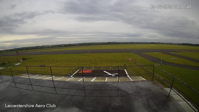 An outdoor view of Leicesterhire Aero Club shows a cloudy sky over a grassy airfield with runways and taxiways in the distance. In the foreground, there is a platform with a railing, a helipad marked with an H and a red square, and some equipment. A windsock is visible on the left side, nearby a small picnic table and a bench. The timestamp on the top right indicates the date as May 6, 2025, at 9:00 PM.