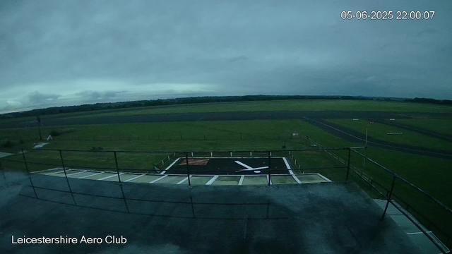 An empty helicopter landing pad with black markings and white lines, surrounded by green grass and open skies with dark, overcast clouds. There are distant fields and a horizon line, with a fence and some equipment visible on the perimeter. The image has a timestamp in the upper right corner and the words "Leicestershire Aero Club" at the bottom left.