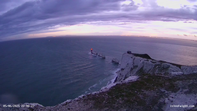 A panoramic view of a rugged white chalk cliff overlooking the sea at sunset or dusk. The sky is mostly overcast with dark clouds, with a hint of orange near the horizon. A narrow pathway runs along the edge of the cliff, with a small building or structure visible near the right side. The ocean has gentle waves, and a lighthouse with a bright red light is situated on a small rock formation just offshore, with sailboats visible in the distance. The timestamp reads "05/06/2025 22:00:27" and the website "isleofwight.com" is displayed in the lower right corner.