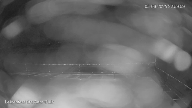 A black-and-white webcam image showing an indoor setting, possibly a sports or recreational facility. There are blurred objects in the foreground and what appears to be a railing or barrier along the bottom of the image. The top right corner displays a date and time stamp: "05-06-2025 22:59:59," and the bottom left corner has the text "Leicestershire Aero Club."
