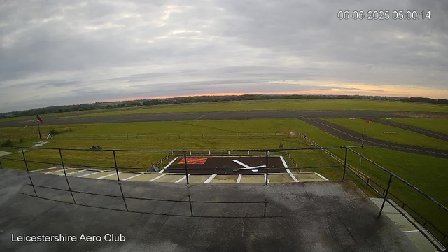 A view from a webcam at Leicesterhire Aero Club showing an empty parking area and the airstrip in the distance under a cloudy sky. The time and date stamp reads 06-06-2025 05:00:14.