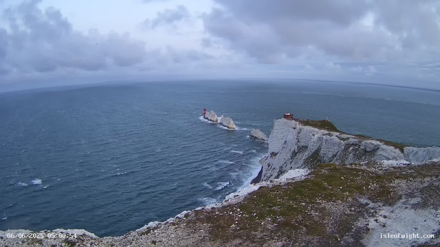 A view of a rugged coastal landscape with white chalk cliffs and a narrow strip of land extending into the sea. There is a small building or hut atop the cliffs, and a lighthouse on a nearby rock formation in the water. The ocean appears vast with gentle waves, and the sky is partly cloudy with patches of blue.