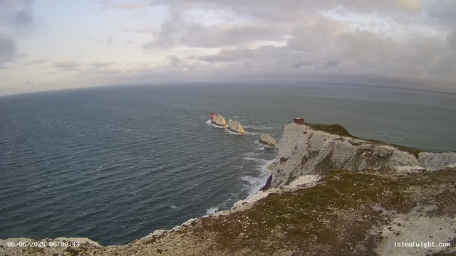 The image shows a viewpoint from a cliff overlooking the ocean. In the distance, there are several large white rock formations with a red and white lighthouse near the second rock. The sky is cloudy, and the time stamp in the lower left corner reads "06/06/2025 06:00:43."