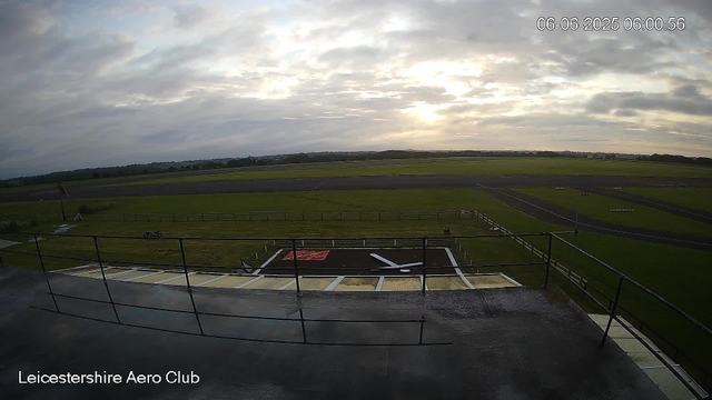 A wide view of the runway and surrounding grassy areas at Leicestershire Aero Club during cloudy weather with the sun partially obscured by clouds, and the timestamp showing June 6, 2025, at 6:00 AM. The image is taken from an elevated position with a railing visible in the foreground.