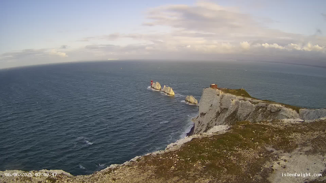 Aerial view of a coastal cliff with a small building on top, overlooking the ocean. Multiple large rocks are visible in the water near the shoreline, and a red and white lighthouse is situated on one of the rocks offshore. The sky is partly cloudy with some sunlight breaking through. The scene is calm and scenic, with the timestamp "06/06/2025 07:00:47" in the lower left corner. The website "isleofwight.com" is printed in the bottom right corner.