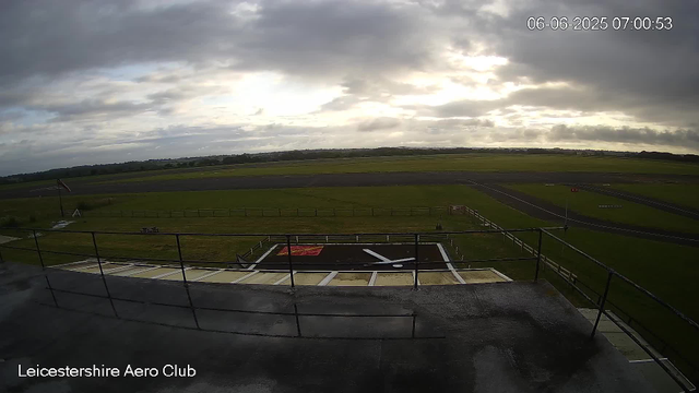 A view from a webcam at Leicesterhire Aero Club shows a cloudy sky above an empty grass airfield. The foreground includes a platform with black and white markings, a red square, and a white arrow. The airfield has runways and taxiways stretching out into the distance, with a fence line separating the grassy areas from the paved surfaces. The timestamp on the image indicates it was taken on June 6, 2025, at 7:00 AM.
