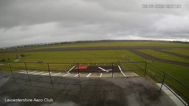A view from a webcam at Leicestershire Aero Club shows an overcast sky with grey clouds. In the foreground, there is a wet tarmac area with a helipad marked by a red square and a white arrow. A black metal railing surrounds the area, and a flagpole with a British flag is positioned on a grassy section to the left. The grassy fields extend into the distance, with a few trees scattered along the horizon and a series of paved runways or taxiways. The scene has a moody, cloudy atmosphere with no visible aircraft or people. The timestamp on the image reads 06-06-2025 08:01:01.