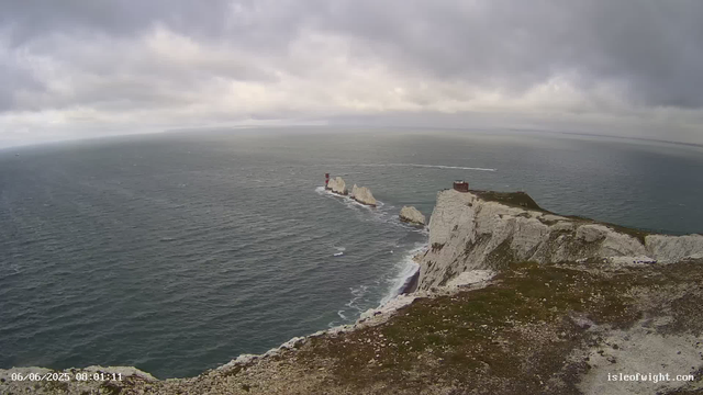 A view of a rugged coastline with tall white chalk cliffs extending into the ocean. On top of the cliffs, there are a few small structures and a lighthouse positioned near the edge. The sea below is choppy with visible waves, and the sky above is overcast with gray clouds. In the distance, the horizon separates the sky from the water. The timestamp in the bottom left corner reads "06/06/2025 08:01:11," and the URL "isleofwight.com" is visible in the bottom right corner.