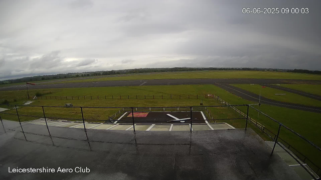 An outdoor view of Leicester Aeroclub's tarmac and runway on a cloudy day. The foreground shows a black helipad with white markings and a red square, with a white pole and flag nearby. Beyond the helipad, there is a grassy area with a small picnic table, a wind sock, and a fence. The runway stretches into the distance, with a line of trees and hills on the horizon under dark, cloudy skies. The date and time displayed in the top right corner are June 6, 2025, at 09:00:03.