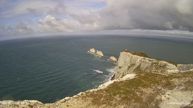 An aerial view of white chalk cliffs and rocky outcrops extending into the ocean, with a red and white lighthouse on one of the rocks. The sky is partly cloudy with patches of blue, and the sea has gentle waves. A small structure, possibly a camera or observation post, is visible on the edge of the cliff. The photo date and time are displayed in the bottom left corner.