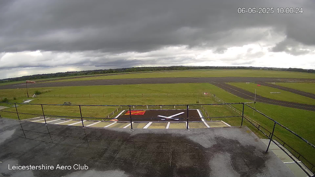 A view from a webcam at Leicestershire Aero Club shows an overcast sky with dark clouds. In the foreground, there is a tarmac rooftop with a wet surface, and a railing surrounds the ledge. Below, a grassy area with a large airplane logo painted on the ground and a red and white windsock pole nearby. Further away, there is a runway and taxiway, with some small signs and markings visible. The surrounding landscape includes expansive green fields and a distant treeline. The date and time in the top right corner indicate it is June 6, 2025, at 10:00 AM.