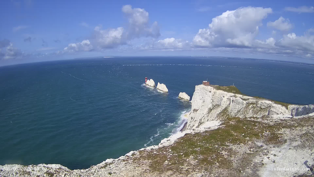 Aerial view of white chalk cliffs along a coastline with a lighthouse on a small peninsula. The sea is deep blue with gentle waves, and a cloudy sky overhead.