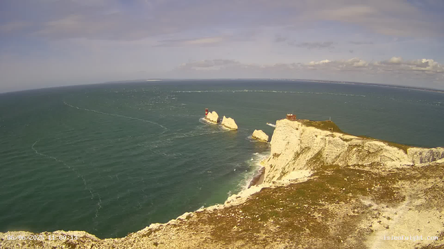 A view of white chalk cliffs along the coastline with a lighthouse on a prominent rock outcrop. The ocean is calm, with waves gently hitting the base of the cliffs, and a partly cloudy sky overhead.