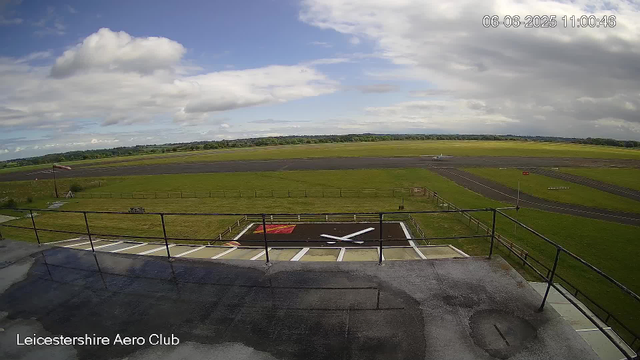 An outdoor view from a webcam at Leicestershire Aero Club showing a runway and surrounding grassy areas under a partly cloudy sky. There is a helicopter landing pad in the foreground with painted helipad markings and a windsock nearby. The date and time stamp in the top right corner reads "06-06-2025 11:00:46".