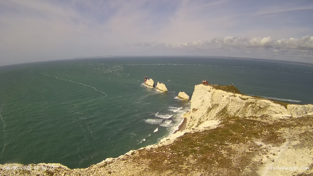 An aerial view of white chalk cliffs along a coastline with a lighthouse on one of the cliffs. The ocean extends into the distance with boat wakes and spray visible, under a partly cloudy sky.