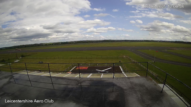 A view from a webcam at Leicestershire Aero Club showing an outdoor scene with a runway in the background, a grassy area with a windsock, a helipad with markings, and a cloudy sky overhead. The image is timestamped June 6, 2025, at 12:00:58.