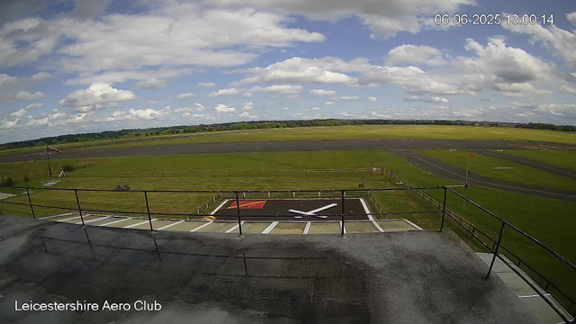 A scenic view of an airfield under a partly cloudy sky. The foreground shows a small helipad with a large white "H" and painted lines. A fence surrounds the airfield, and there is a flagpole with a flag near the helipad. In the background, the runway and taxiways stretch across the landscape, with green fields beyond. The date and time stamp in the top right corner shows "06-06-2025 13:00:14". The bottom left corner has the text "Leicestershire Aero Club."