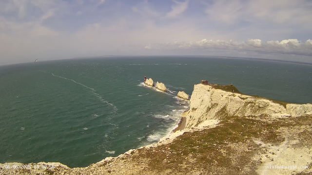 An aerial view of white chalk cliffs along the coastline, with a lighthouse on a small rocky islet in the sea. The cliffs extend into the distance, meeting a partly cloudy sky, and a boat is visible far out on the water. The date and time are displayed in the bottom left corner.