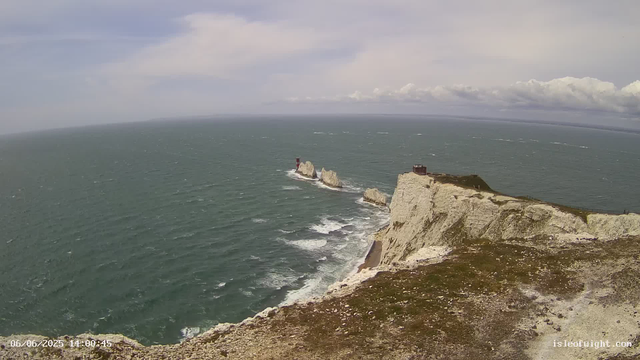 A view of rugged white chalk cliffs extending into the ocean with several prominent sea stacks. The cliffs are topped with grassy areas and small structures. The sea is choppy with visible waves, and the sky above is partly cloudy with patches of blue. The timestamp in the lower left corner indicates the date and time as June 6, 2025, at 14:00:45.