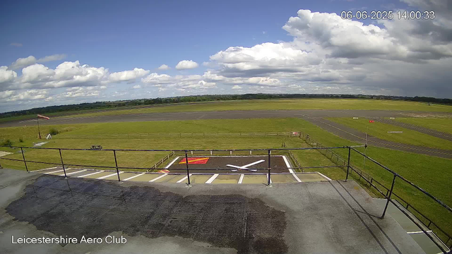 A wide view of the Leicesterhire Aero Club airfield under a partly cloudy sky. The foreground shows a small fenced helipad with red and white markings, and a parking area with white lines. Beyond the helipad, there is a grassy field with a flagpole on the left side. The runway extends into the distance, with several small markers and lights. The horizon features a line of trees and open countryside. The date and time are displayed in the top right corner: June 6, 2025, at 14:00:33.