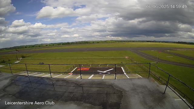 A wide view of a runway and grassy airfield at Leicestershire Aero Club under a partly cloudy sky, with some dark clouds and patches of blue. The image is timestamped June 6, 2025, at 14:59:49. In the foreground, there is a rooftop with safety railings, a helicopter landing pad with a large white "X," and some painted markings. The horizon shows a line of trees and fields in the distance.