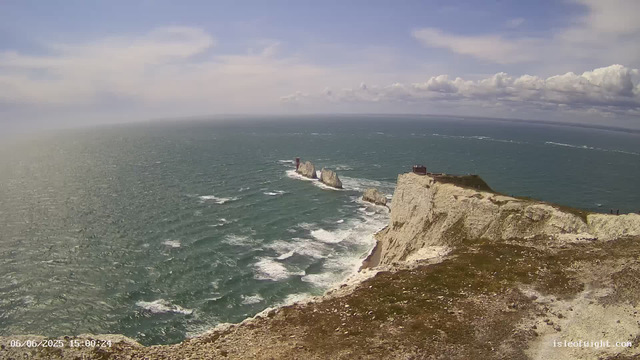 A panoramic view of the ocean with white cliffs and sea stacks, under a partly cloudy sky. The foreground shows the edge of a grassy cliff, and in the distance, there are two small structures on the cliffs. The image has a timestamp of June 6, 2025, at 15:00:24.