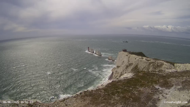 A view of a rugged coastline with white chalk cliffs extending into the ocean. There are three prominent sea stacks near the shore, with a lighthouse on top of one of the stacks. A large ship is visible in the distance on the ocean. The sky is cloudy with patches of light breaking through, and the water has visible waves. The timestamp shows "06/06/2025 16:01:03" and the website "isleofwight.com" is in the bottom right corner.