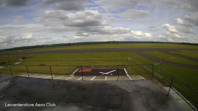 A wide view of an airfield under a partly cloudy sky. The foreground shows a rooftop with a helipad marked with an 'H' and a red square, surrounded by a fence. Behind the helipad, there are grassy areas, taxiways, and runways stretching into the distance with green fields and a treeline on the horizon. The timestamp in the upper right corner reads "06-06-2025 16:00:59," and the bottom left corner displays "Leicestershire Aero Club."