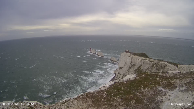 A view of white chalk cliffs extending into the ocean under a cloudy sky. The cliffs have a rugged, steep face with patches of green grass on top. In the distance, there are two small lighthouse-like structures on the rocks protruding from the water. The sea appears choppy with white waves crashing against the base of the cliffs. The image timestamp shows "06/06/2025 17:00:42" and the website "isleofwight.com" is visible at the bottom right corner.