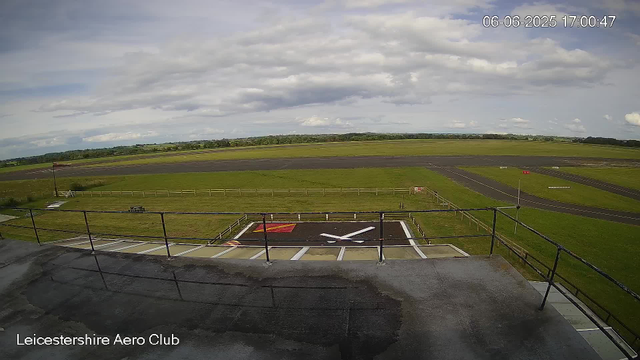 An outdoor view of Leicesterhire Aero Club showing an airfield with a runway and grassy areas. A landing pad with a white helicopter symbol and colored markings is visible in the foreground. The sky is partly cloudy, and the scene is calm and open, with distant trees on the horizon. The date and time can be seen at the top right corner: 06-06-2025 17:00:47.