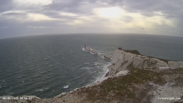 A view of the ocean with white chalk cliffs extending into the sea. There are several rock formations in the water, including a tall, narrow lighthouse on one of the rocks. The sky is partly cloudy with the sun breaking through the clouds, casting a soft light over the scene. The timestamp indicates the date and time as June 6, 2025, at 6:00 PM.