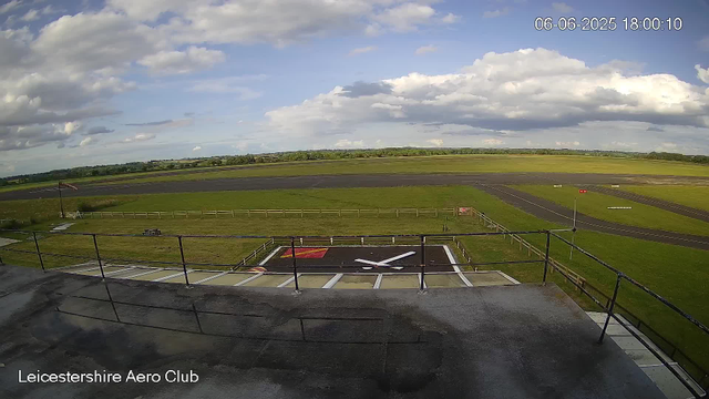 A view of an aircraft tarmac and surrounding grassy fields under a partly cloudy sky. There is a large painted clock with white hands and a red background on the ground in the foreground, and a small windsock on a pole to the left. The scene is labeled "Leicestershire Aero Club" at the bottom left corner, and the top right corner displays the date and time: 06-06-2025, 18:00:10.