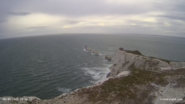 A view of white chalk cliffs overlooking the ocean under a cloudy sky. The cliffs extend into the sea, with a notable lighthouse on a small island just off the coast. The landscape appears rugged, with some patches of grass and rocky surfaces on the cliff top. The image has a timestamp of June 6, 2025, at 19:00:55.