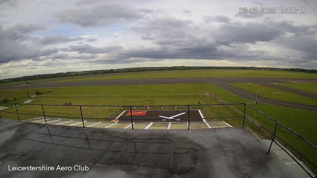 A view from a webcam at Leicestershire Aero Club shows an overcast sky with dark clouds. In the foreground, there is a concrete balcony with a black metal railing. Below, a small helipad marked with a large white "H" and a red border is visible. Beyond the helipad, there is a fenced grassy area with a windsock on a pole to the left. Further in the distance, the airfield features a runway and taxiways, surrounded by expansive green fields and a distant treeline. The timestamp on the image indicates it was taken on June 6, 2025, at 19:00:48.