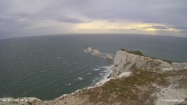 A view of the white chalk cliffs of Étretat, France, with a lighthouse on the edge of the cliffs. The ocean surrounds the cliffs, and the sky is cloudy with a hint of sunlight breaking through. The overall scene appears to be taken during late evening or early night.