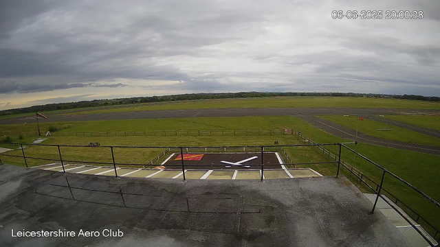 An outdoor view from a webcam at Leicester Aeroclub shows a cloudy sky with dark gray clouds. In the foreground, there is a helicopter landing pad marked with a large white "H" on a red background, surrounded by a small fence and accessible from a balcony or viewing area with a metal railing. The grass around the landing pad is green, and in the background, there is a paved runway stretching across an open field. The scene appears calm and overcast. The timestamp indicates the date and time as June 6, 2025, at 8:00 PM.