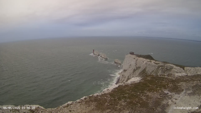 A view of the white chalk cliffs of Dover, England, with the sea stretching out beyond. The cliffs feature two small structures at the top and a lighthouse on one of the distant rock formations. The sky is cloudy with hints of light, suggesting an early evening or late afternoon setting. The image is timestamped "06/06/2025 21:00:08" and has the website "isleofwight.com" in the corner.