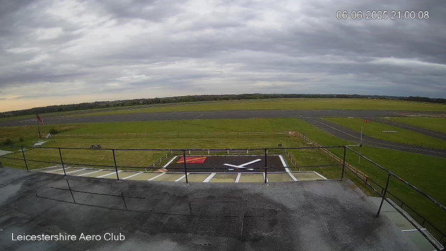 A view from a webcam at Leicestershire Aero Club shows an overcast sky with dark clouds. In the foreground, there is a runway and a small helipad with a white helicopter silhouette painted on it. The helipad is surrounded by a safety fence. To the left, there is a flagpole with a flag, and a small bench nearby. The grassy area is well-maintained, and the overall scene appears calm with no aircraft or people visible. The date and time in the upper right corner are June 6, 2025, at 21:00:08.