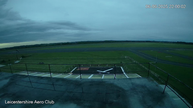 The image shows an outdoor view of a landing pad and surrounding grassy area at Leicester Aero Club. The sky is overcast with dark clouds, indicating cloudy or potentially rainy weather. The landing pad has a large white "X" painted on it, and there is a small structure with a railing in the foreground. In the distance, there are open fields, a track or runway, and a wind turbine visible on the left side. The time and date stamp in the top right corner indicates it was captured on June 6, 2025, at 10:00 PM.