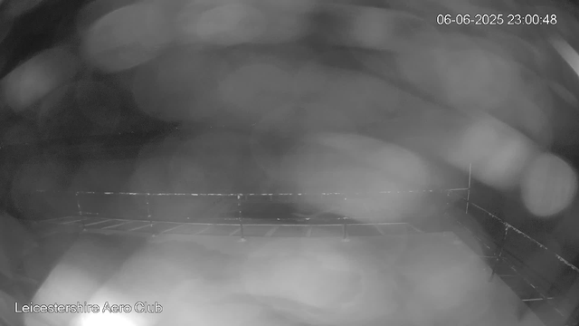 This is a black-and-white image from a webcam, showing a view of a rooftop or outdoor area at night. There are some faint structures or barriers along the edges, possibly a fence or railing, and the sky appears dark with some blurry light or reflections. The date and time in the upper right corner indicate it was captured on June 6, 2025, at 11:00 PM. The text in the lower left corner reads "Leicestershire Aero Club."