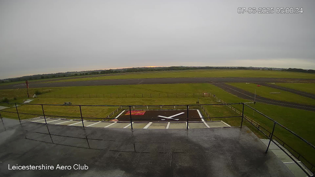 A view from a webcam at Leicestershire Aero Club shows an overcast sky above an airfield with a runway in the background. In the foreground, there is a landing pad with a large clock painted on it, bordered by a black railing. The date and time stamp in the upper right corner reads "07-06-2025 05:00:34."