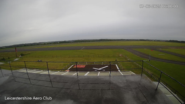 A large grassy area surrounds a runway at Leicestershire Aero Club. In the foreground, there is a helipad with a white helicopter symbol. The weather appears overcast with gray clouds. The landscape is flat, with distant trees and fields visible beyond the runway. The timestamp shows the date as July 6, 2025, at 6:00 AM.