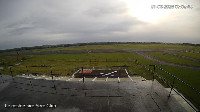 A view from a webcam at Leicesterhire Aero Club showing an overcast sky with the sun partially obscured by clouds. The scene includes a runway, grassy areas, and a small helipad in the foreground. The date and time stamp in the top right corner reads "07-06-2025 07:00:43."