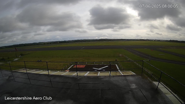 An overcast sky with dark, heavy clouds over a large open airfield at Leicestershire Aero Club. The image shows a concrete observation area with a railing in the foreground, and a clock and "Leicestershire Aero Club" label are visible. There is a runway extending into the distance, surrounded by green fields, with a windsock on the left side of the image. The runway appears wet and the weather looks cloudy and gloomy.