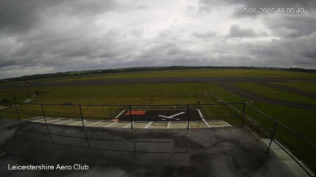 An overcast sky with dark, heavy clouds over a small airport runway. In the foreground, there is a rooftop or observation deck with a black safety railing. A large black and white helicopter landing pad with a prominent white "X" in the center is visible. The surrounding grassy area is green and well-maintained, and an American flag is flying on a flagpole near the left side of the image. The background shows open fields and distant trees. The timestamp indicates the date and time as July 6, 2025, at 10:00 AM. The caption "Leicestershire Aero Club" is displayed in the bottom left corner.
