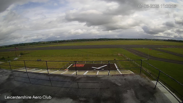 An empty airport runway and taxiway scene viewed from a webcam at Leicestershire Aero Club. The sky is partly cloudy with patches of sunlight, and the runway is marked with various lines and numbers. There is a windsock on the left side, and the surrounding grassy areas extend into a distant horizon. The timestamp indicates the date as July 6, 2025, at 11:00 AM.