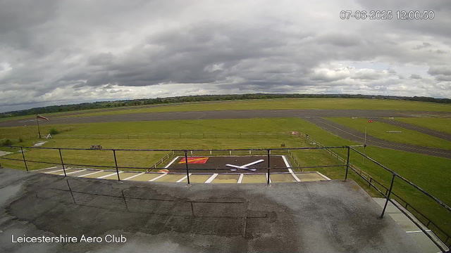 A view from a webcam at Leicestershire Aero Club showing an empty aircraft parking area with a helicopter landing pad marked with a white "X" in the foreground. Beyond the parking area, there is a grassy field leading to a runway, with overcast skies and dark clouds overhead. The scene appears calm with no aircraft or people visible.