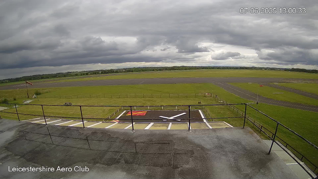 The image shows an overcast sky with dark clouds above a small airport runway. In the foreground, there is a helicopter landing pad with a white and red markings and a large white "X" in the center. The boundary fence surrounds a grassy area, and a windsock is visible on the left side. The runway extends into the distance with some markings and runway lights. The scene is from a webcam at Leicestershire Aero Club, with the date and time displayed as July 6, 2025, 13:00:33.